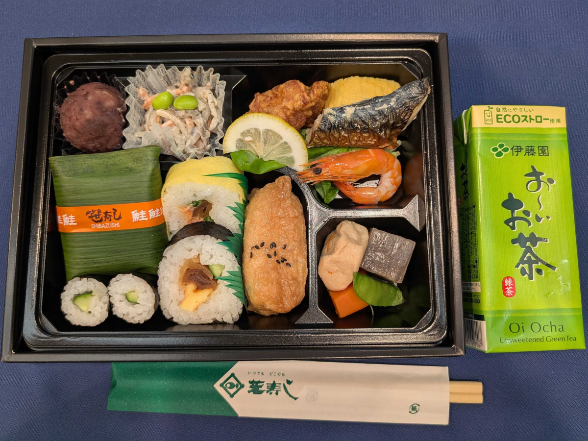さまざまな寿司やおかずが詰まった和風弁当の上からの写真。笹寿司や巻き寿司、いなり寿司、焼き魚、海老、煮物、きんぴら風の和え物、あんこのおはぎが仕切りの中に並び、右側に紙パックの  お～いお茶  と箸袋が添えられている。