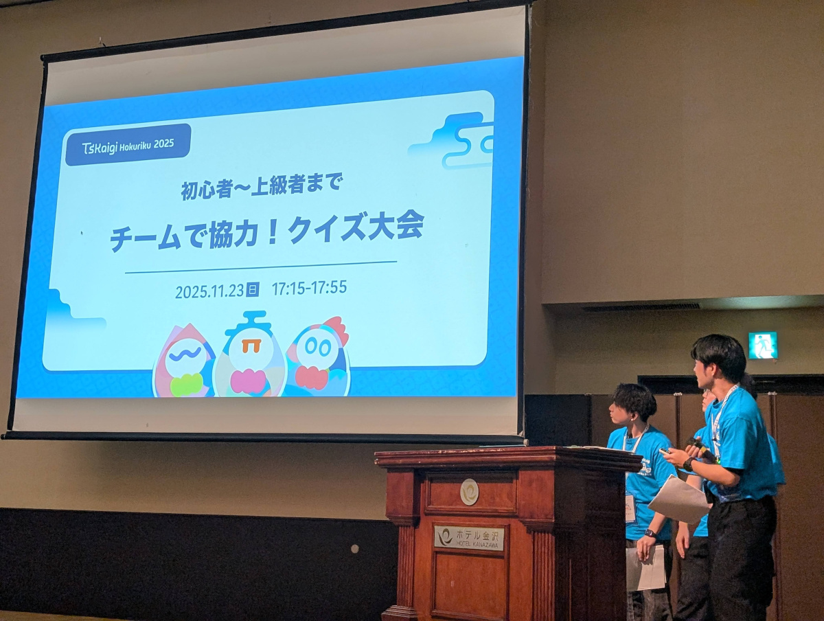 カンファレンス会場の壇上。スクリーンに「初心者〜上級者まで チームで協力！クイズ大会」「2025.11.23」などの文字とキャラクターイラストが表示され、手前に青いスタッフTシャツを着た登壇者たちが立っている。