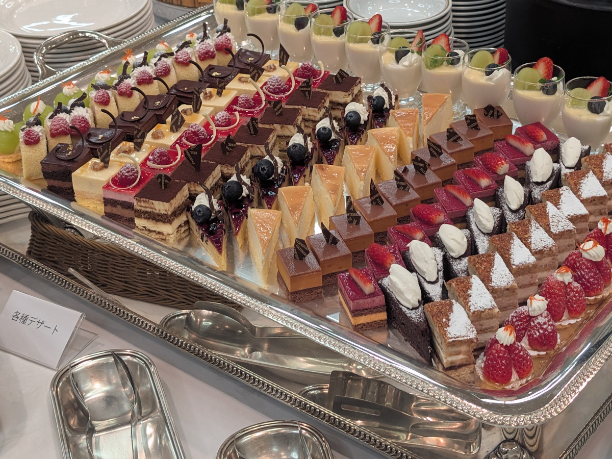 大きな銀色トレーに一口サイズの色とりどりのケーキが整然と並ぶデザートビュッフェ。ベリーやチョコレートが乗ったケーキの列の奥に、フルーツを乗せたグラスデザートが並び、手前に取り分け用トングと「各種デザート」の札が置かれている。