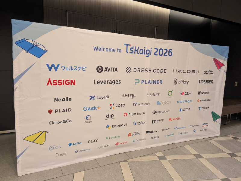 『Welcome to TsKaigi 2026』と書かれたカンファレンスの巨大なスポンサーボード。カラフルな紙飛行機のイラストとともに、多数の協賛企業のロゴが並んで印刷されている。