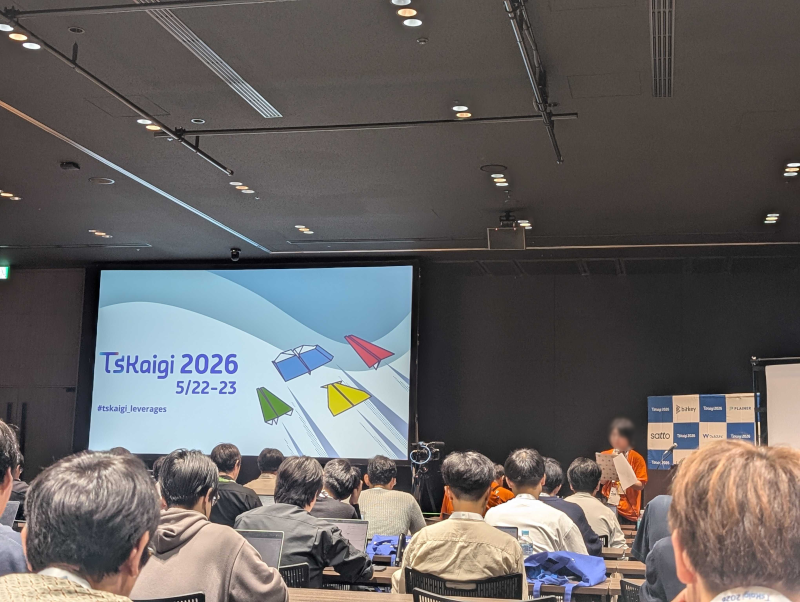 『TsKaigi 2026 5/22-23』と表示された大きなスクリーンに向かって、多数の参加者が着席しているカンファレンス会場の様子。右前方ではスタッフがマイクの前に立って話をしている。