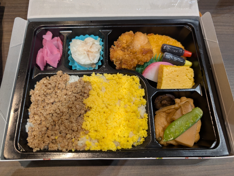 ご飯の上に鶏そぼろと炒り卵が乗ったそぼろ弁当。シュウマイ、唐揚げ、コロッケ、玉子焼き、かまぼこ、煮物（椎茸、筍、さやえんどう）、ピンク色の漬物など、豊富なおかずが黒い仕切り付きの容器に詰められている。