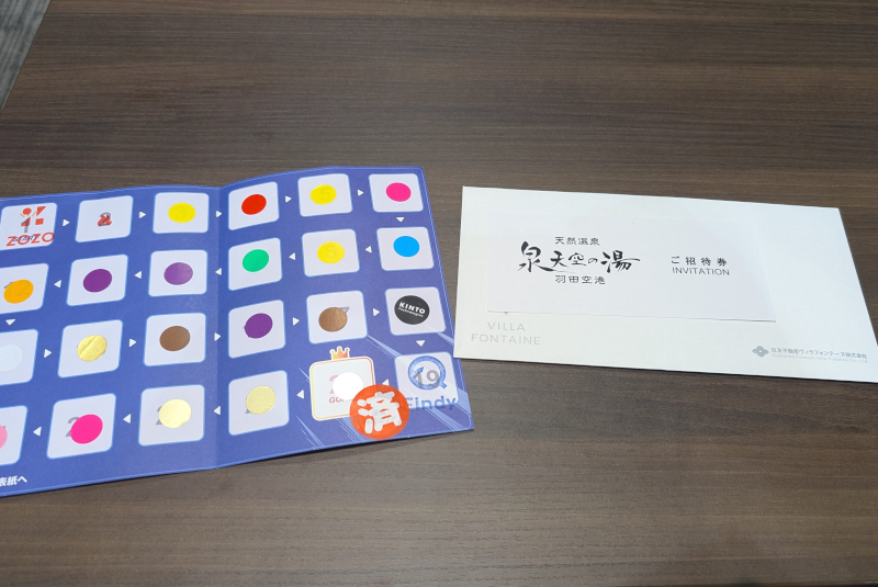 木目の机の上に置かれたスタンプラリーの台紙と招待券。左側の台紙には企業ロゴの上に様々な色の丸いシールが貼られており、右側には『天然温泉 泉天空の湯 羽田空港 ご招待券 INVITATION』と書かれた白い封筒とチケットが置かれている。