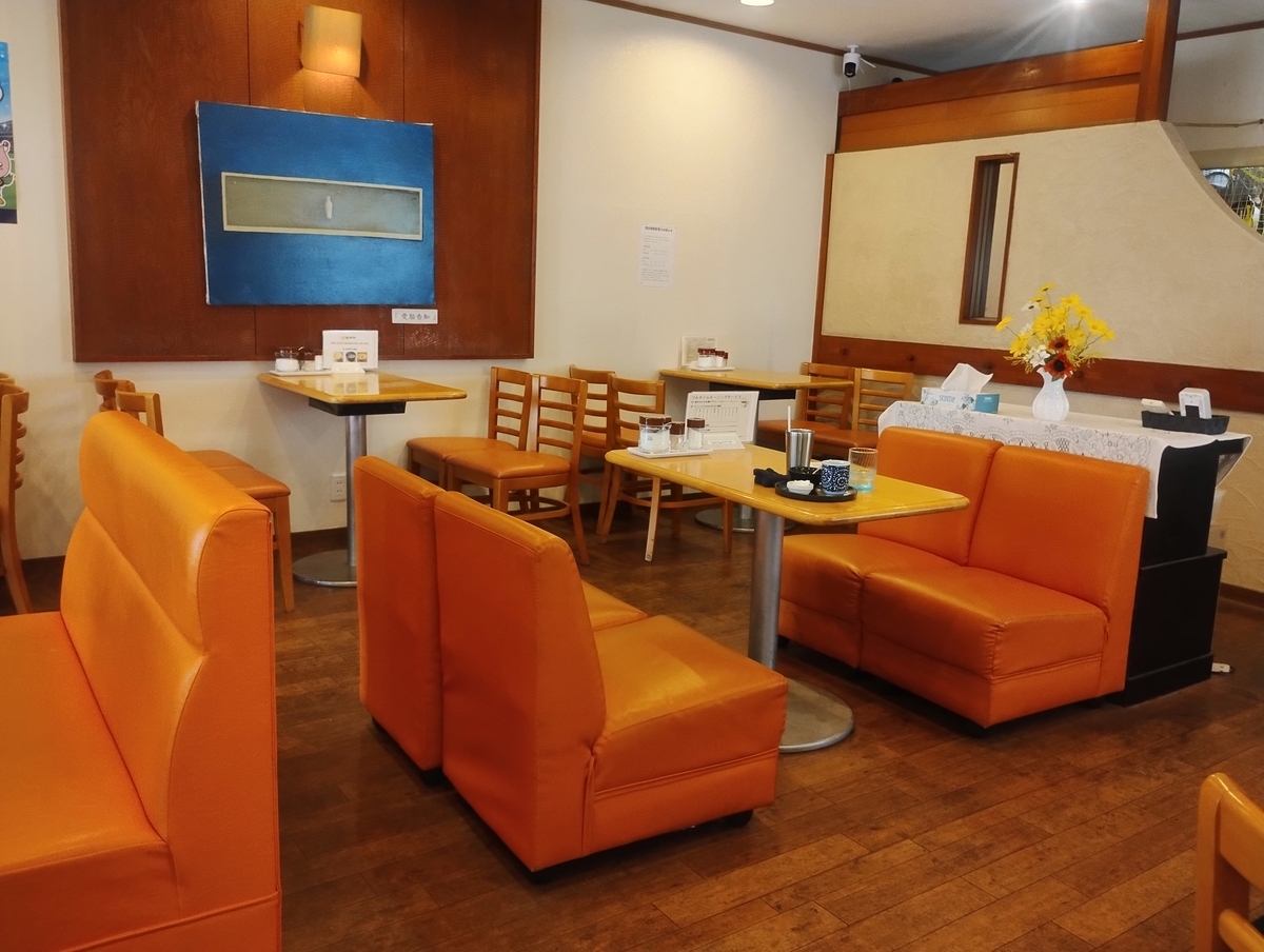 喫茶店『マルミツ』の店内。オレンジ色のソファ席と木製の椅子が並び、落ち着いた雰囲気の空間。壁には青い絵画が飾られ、テーブルには飲み物や調味料が置かれている。