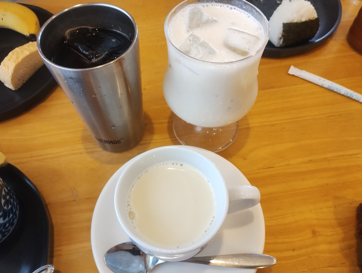 喫茶店『マルミツ』のテーブルに並ぶドリンク。ステンレス製タンブラーのアイスコーヒー、グラス入りの豆乳ドリンク、白いカップのホットミルクティーが置かれ、奥にはおにぎりやバナナなどのモーニングセットが見える。