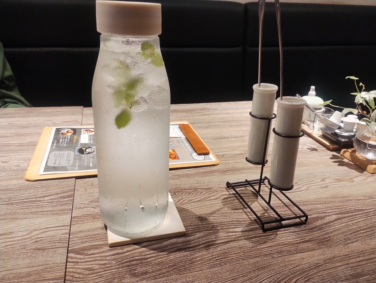 ミントの葉が入ったガラスボトルの水と、細長い白いドレッシングボトルが並ぶテーブル。木目のテーブルにメニューが置かれた、おしゃれなカフェの雰囲気。
