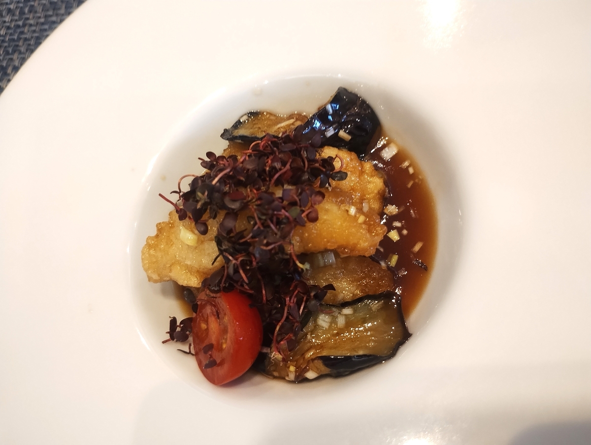 甘酢ソースが絡んだ白身魚のフリットと素揚げしたナス、ミニトマトに、スプラウト(芽ジソ)がトッピングされ、中央が窪んだ白い大皿に盛り付けられた料理