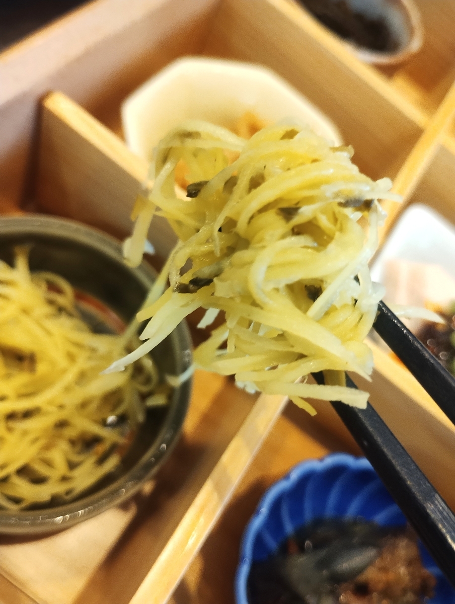 千切りのジャガイモと細かく刻んだ高菜の和え物を、黒い箸で持ち上げ、カメラに近づけている様子。背景には、木箱に入った他の小鉢や、テーブルの上に並べられた様々な前菜(海藻、卵料理、肉料理など)が、ぼんやりと写っている。