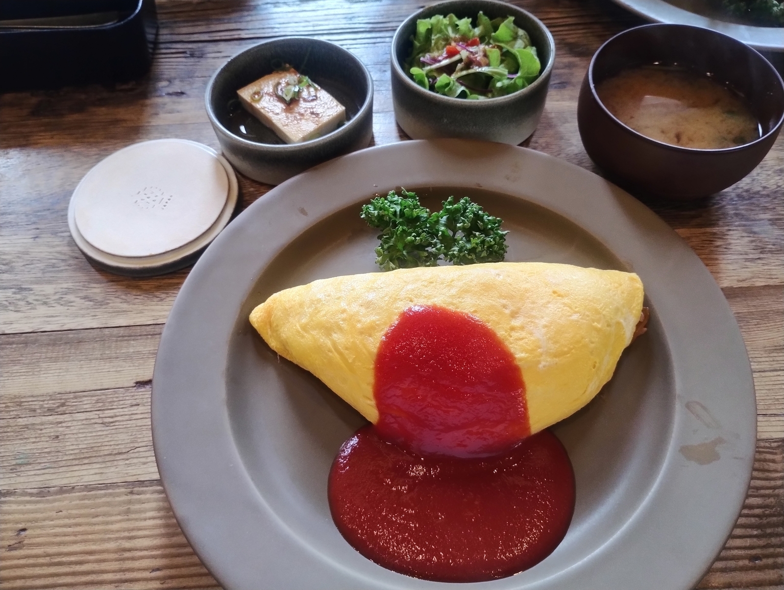 オムライスの定食。黄色い卵に包まれたオムライスの上にケチャップがかかり、横にパセリが添えられている。奥には冷ややっこ、サラダ、味噌汁が並ぶ