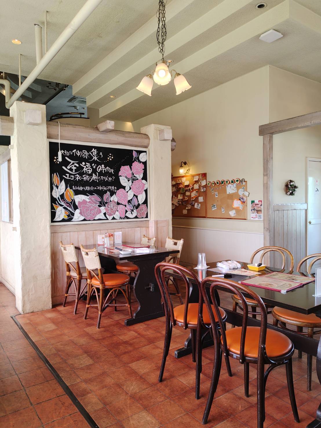 ユコーネの店内。木のテーブルと椅子が並び、黒板に描かれた花のイラストとメッセージが目を引く。奥には掲示板があり、明るい照明が温かみのある雰囲気を作っている。