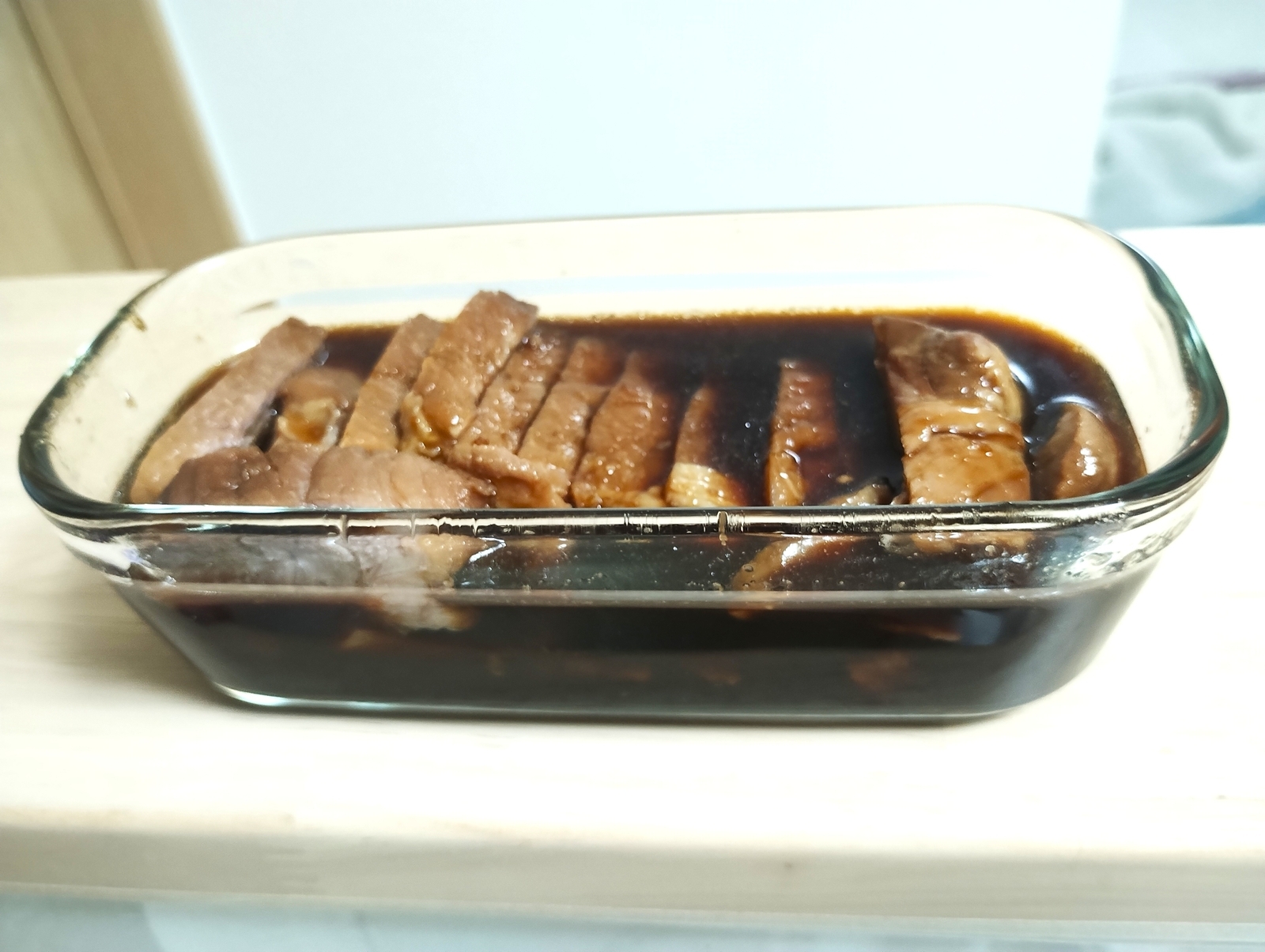 ガラス容器に並べた焼き豚を、濃いタレに漬け込んでいる様子。照りのあるタレが肉にしみ込んでいる。