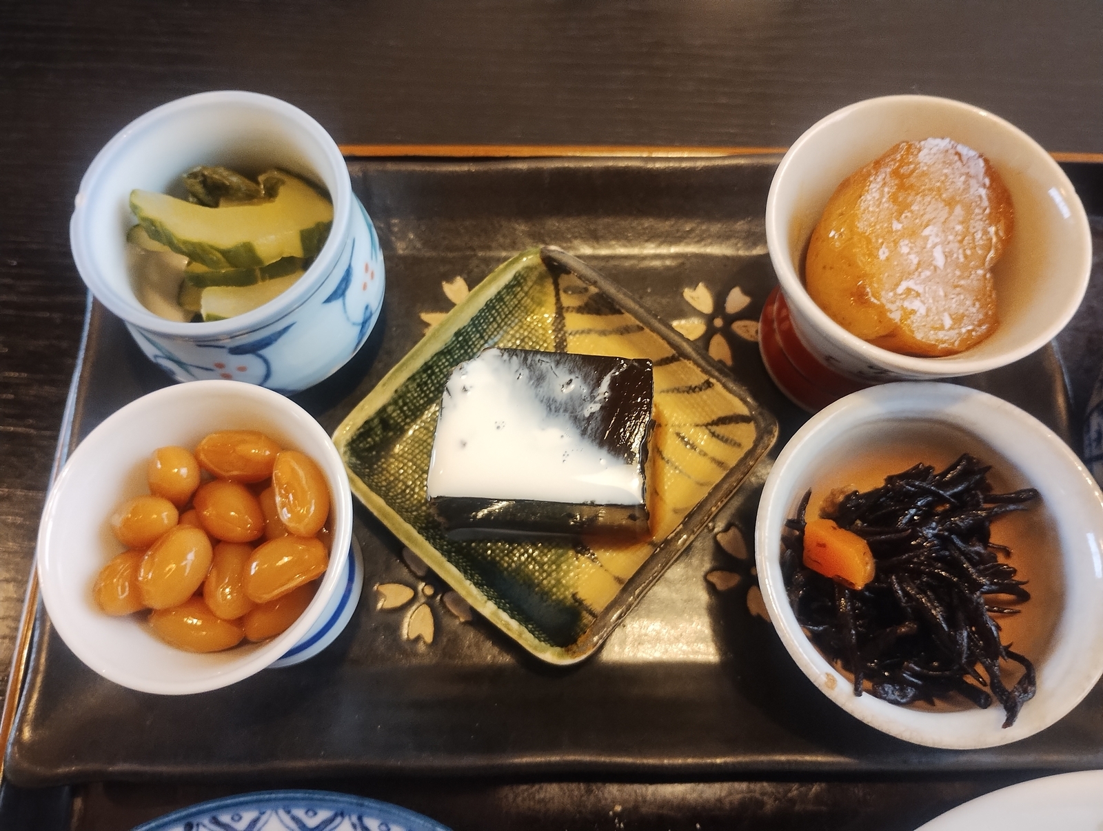 茶色のお盆に、煮豆、キュウリのピクルス、ヒジキ、じゃがいもの煮もの、コーヒー寒天が並ぶ