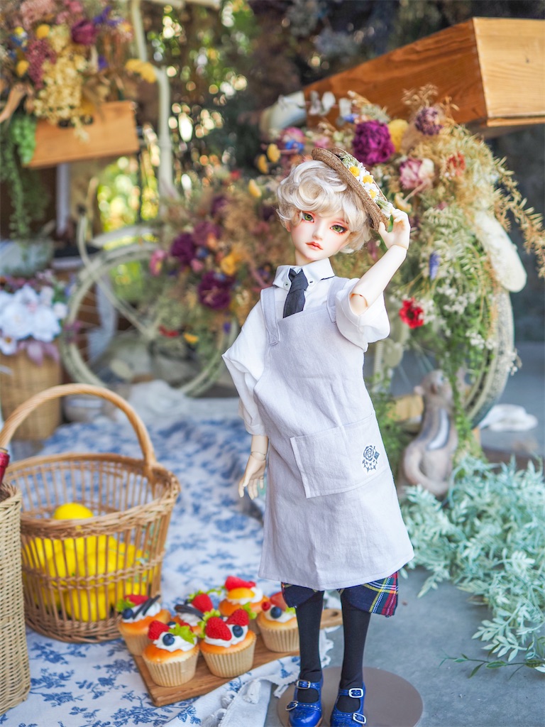 花で飾りつけられた自転車を背景に、半袖半ズボンの学生服にエプロンとストローハットを合わせたつかさくんが立っている写真。ブロンドの短髪で少年らしい雰囲気。