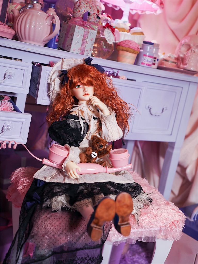 ファンシーなピンクを基調とした部屋の中で、黒いドレスを着た赤毛のつかさくんがテディベアと受話器を膝に載せて座っている写真。
