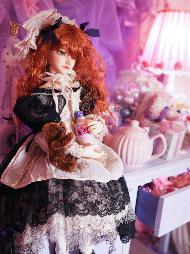 ファンシーなピンクを基調とした部屋で、机の上に座って小瓶を手に取りテディベアと一緒に見つめているつかさくんの写真。