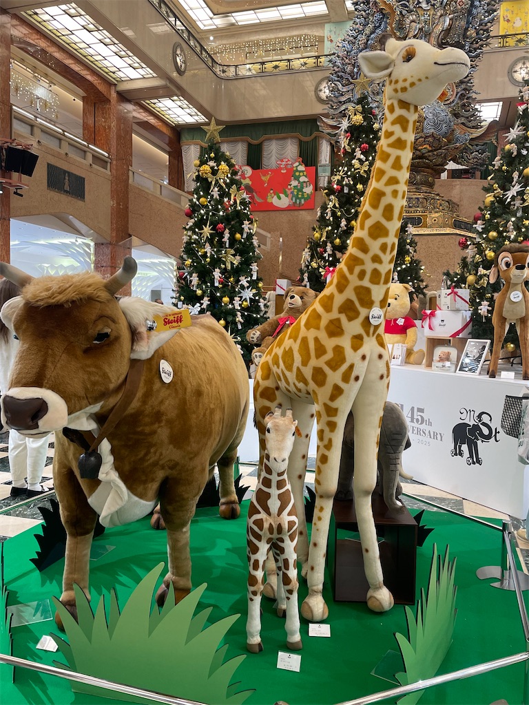 大きいキリンと牛のぬいぐるみの展示の写真。後ろにはテディベアが飾り付けられたクリスマスツリーが写っている。