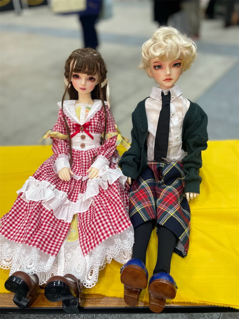 赤いセーラードレスを着た女の子のドールと、学生服姿のつかさくんが一緒に黄色いテーブルの上に座っている写真。左に座っている女の子のドールはあすとらさんのおうちの結衣さん。