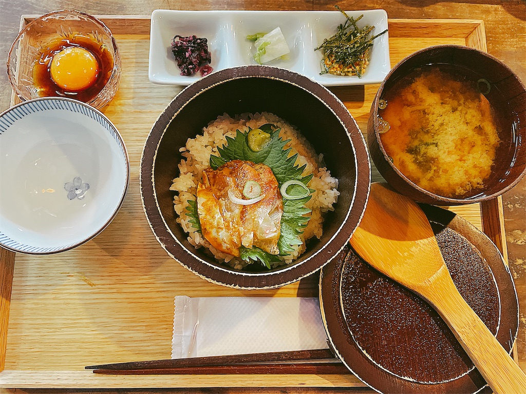 鯛の切り身が載ったおひつのご飯、味噌汁、黄ニラしょうゆがかかった卵の黄身、漬物がお盆の上に載っている、定食の写真。