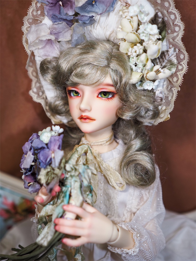 ビオラや鈴蘭、勿忘草の布花で飾られたゴージャスなボンネットを着けたつかさくんの写真。ビオラの花束を持ち、アイボリーのドレスを着て、緑がかったブロンドの巻き髪のウィッグを着けている。