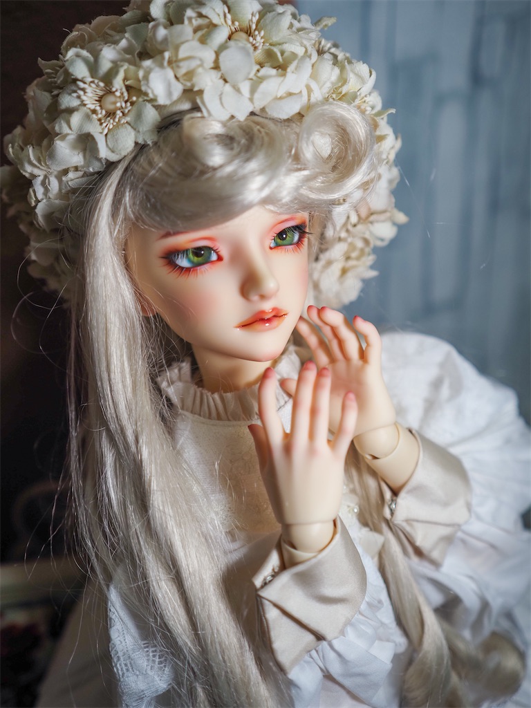白のドレスを着て生成り色の帽子を被ったつかさくんが、顔の近くで両手を合わせている写真。両手のハンドパーツにはメイクがピンクで施されている。