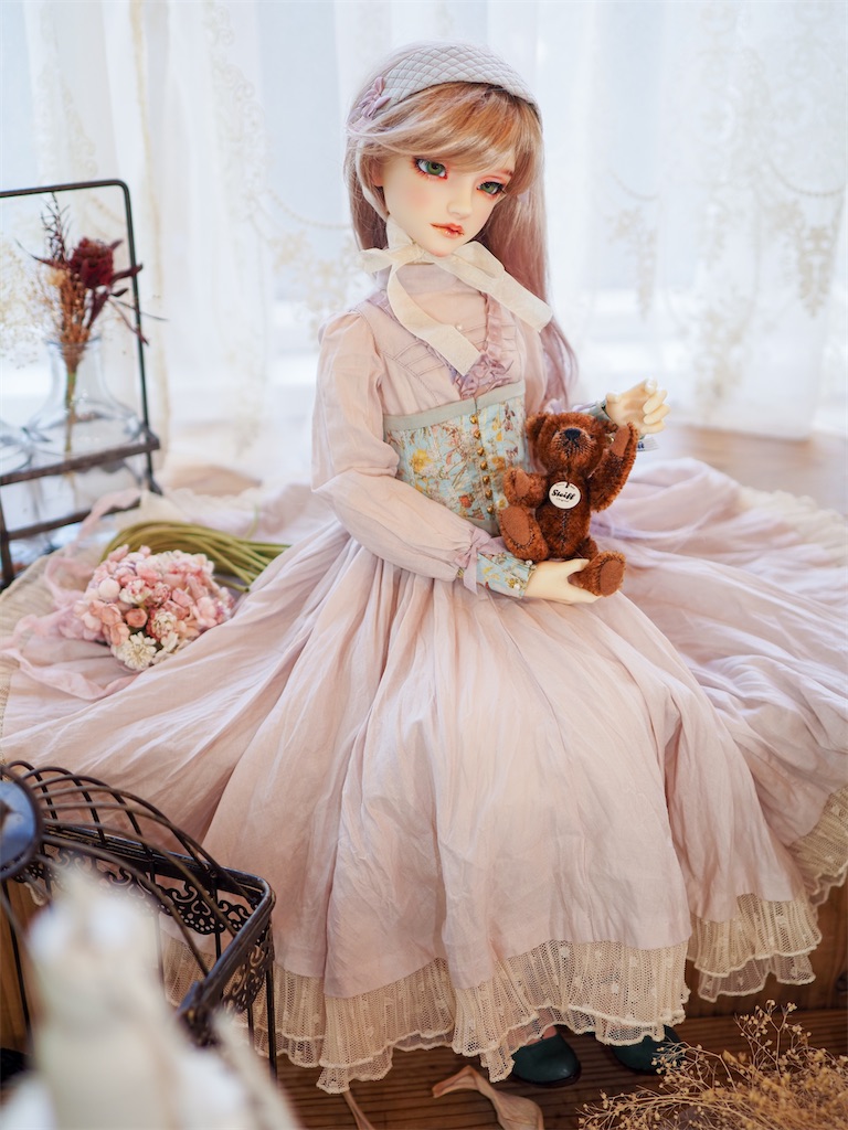 淡いピンクのドレスに水色のコルセットを合わせたコーディネートのつかさくんが座っている写真。ミルクティーブラウンのロングヘアのウィッグを着けている。