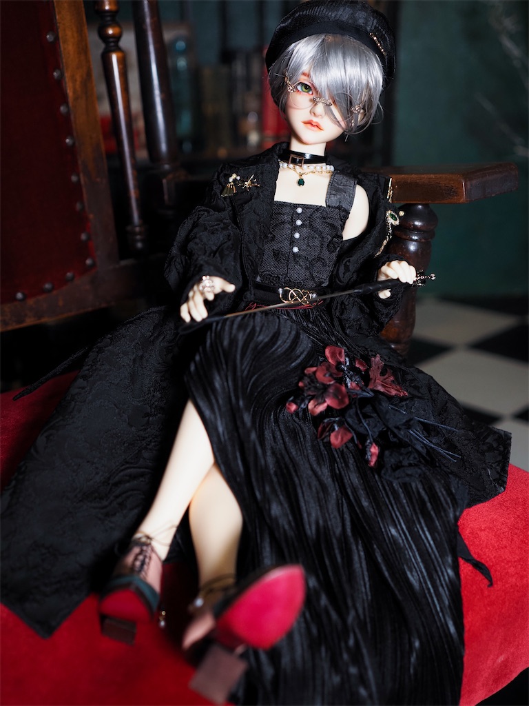 黒いモノトーンのドレスに銀髪のメカクレのショートヘア、シルバーの丸眼鏡、黒い乗馬鞭を合わせて、ピンクとグリーンの革靴を履いたつかさくんの写真。