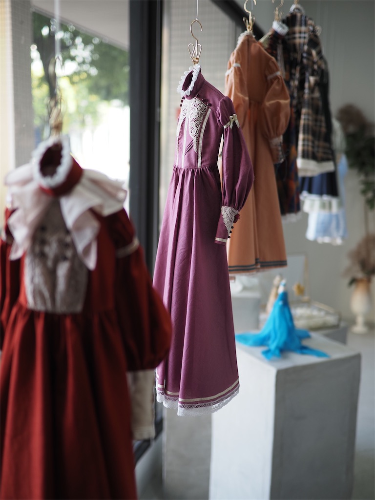 赤やプラムピンク、アプリコットなど様々な色合いのドールサイズのドレスが天井から吊り下げられ、太陽の光を浴びている写真。