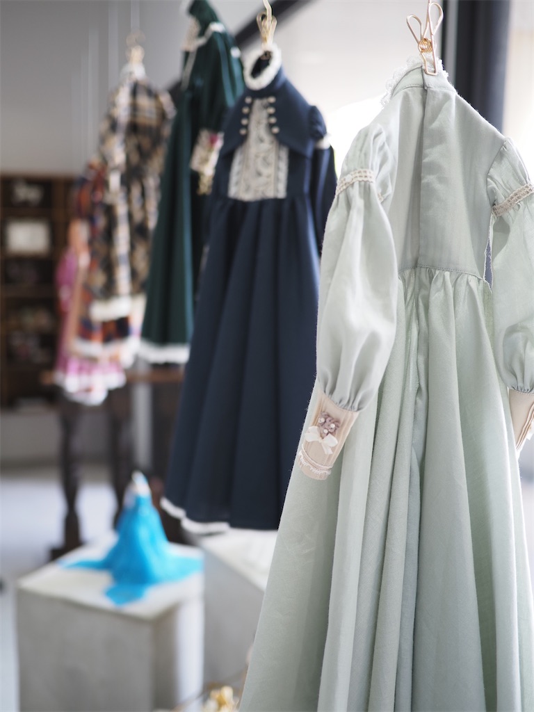 個展の奥から入り口を眺める視点で撮影した写真。大きなドレスが吊り下げられ、小さなドレスは台の上に飾られて、太陽の光を受けている。
