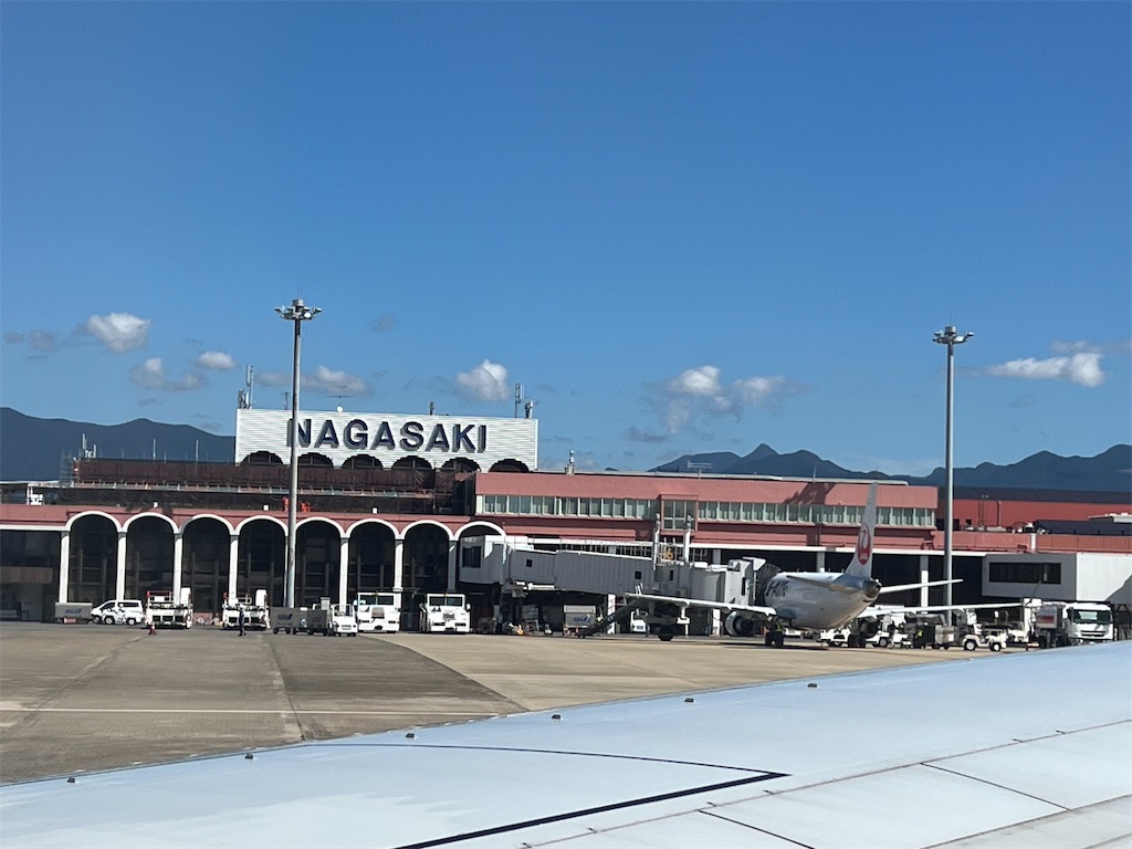 長崎空港。ローマ字でながさきと大きく書かれ、飛行機が一機、作業車っぽいものがたくさん見える。建物の背は低く横長。奥に山も見える。