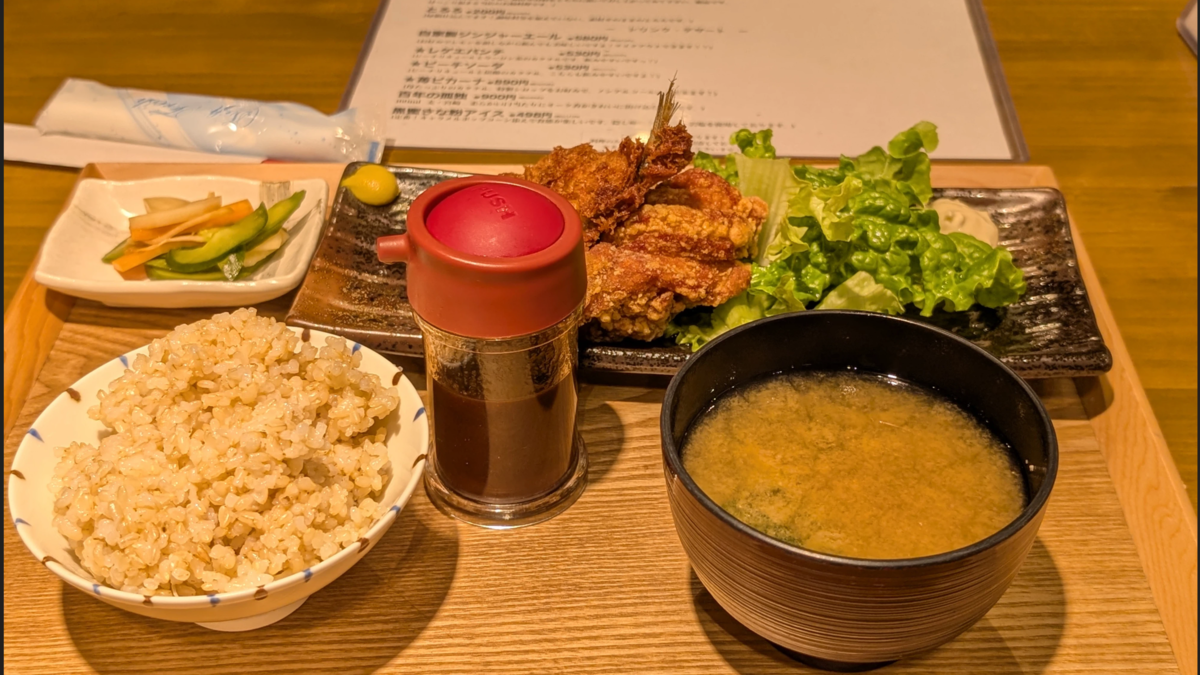 アジフライ&唐揚げ定食。左上から時計回りに、漬け物、アジフライと唐揚げ、味噌汁、ソース瓶、玄米。揚げ物には付け合わせのレタスとからし、タルタルソースも。