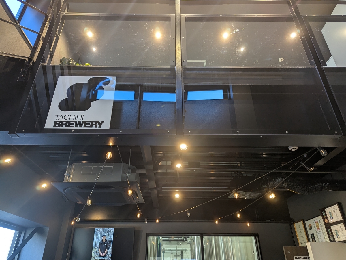 立川・立飛にあるクラフトビール醸造所「TACHIHI BREWERY」の店内。天井の配管や照明、ロゴ看板が見える工業的デザイン空間。