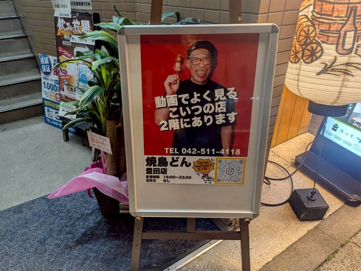 焼鳥どん豊田店の外看板。SNSで話題の日垣さんの写真が大きく掲載されており、『動画でよく見るこいつの店 2階にあります』と書かれた目印の看板。豊田駅北口からすぐのアクセスの良さを示す写真。