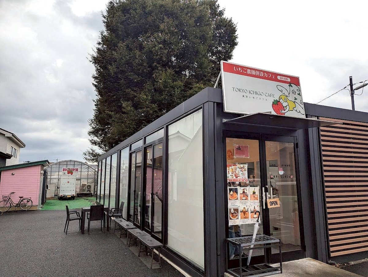 東京いちごカフェの外観。黒いフレームのガラス張りの店舗と、入口上部に「TOKYO ICHIGO CAFE 東京いちごカフェ」と書かれた看板。奥には併設されたいちご農園のビニールハウスがあり、分倍河原駅から徒歩8分の立地にあるカフェ。