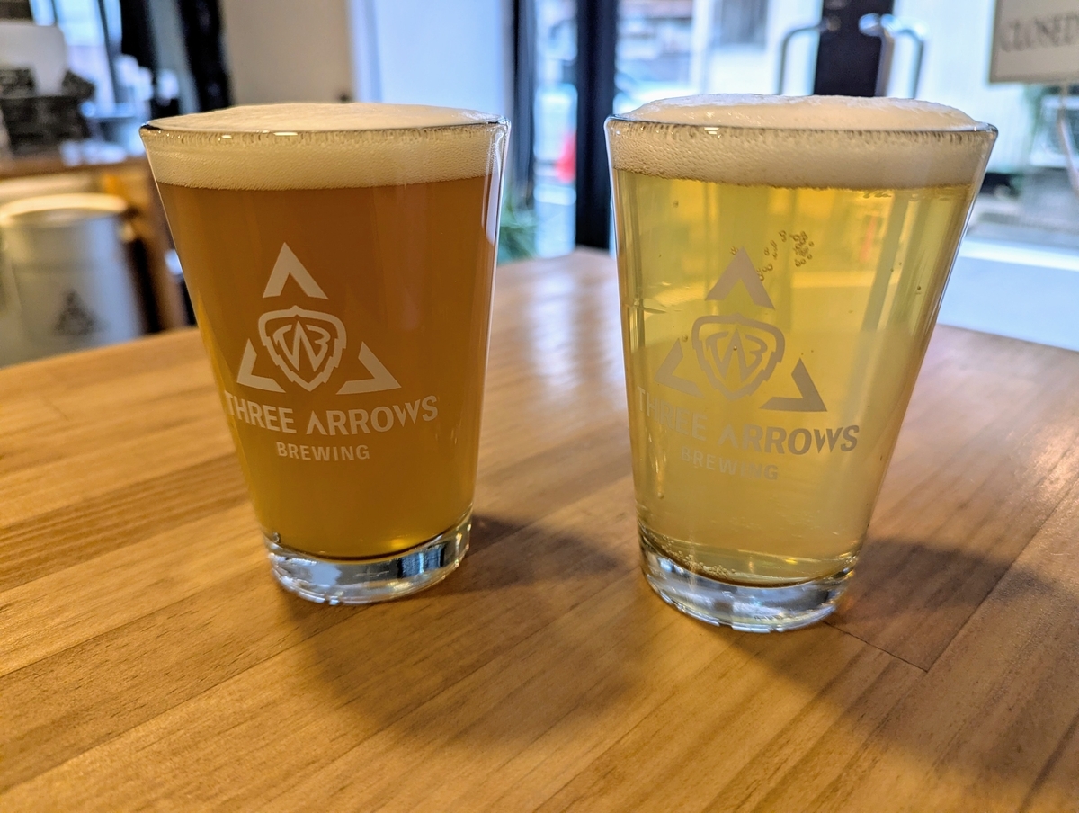 THREE ARROWS BREWINGのロゴ入りグラスに注がれた2杯のクラフトビール。左は濁りのあるオレンジがかった色のダブルIPA、右はクリアな淡いゴールドのコールドIPA。木目のカウンターに並んでいる