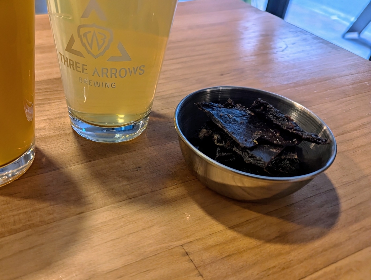 THREE ARROWS BREWINGの自家製鹿ジャーキー。ステンレスの小皿に濃い色のジャーキーが盛られ、隣にロゴ入りグラスのクラフトビールが置かれている