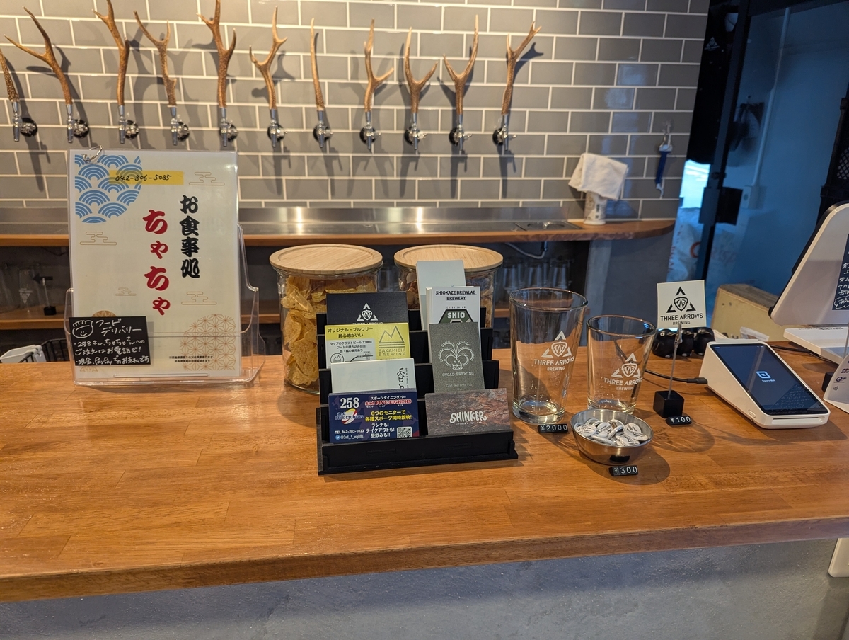 THREE ARROWS BREWINGのカウンター。お食事処ちゃちゃのフードデリバリーメニュー、坂道ブルーイングやSHIOKAZE BREWLABなど他ブルワリーのショップカード、ロゴ入りグラス、キャッシュレス決済端末が並んでいる。奥には鹿の角のビールタップが見える