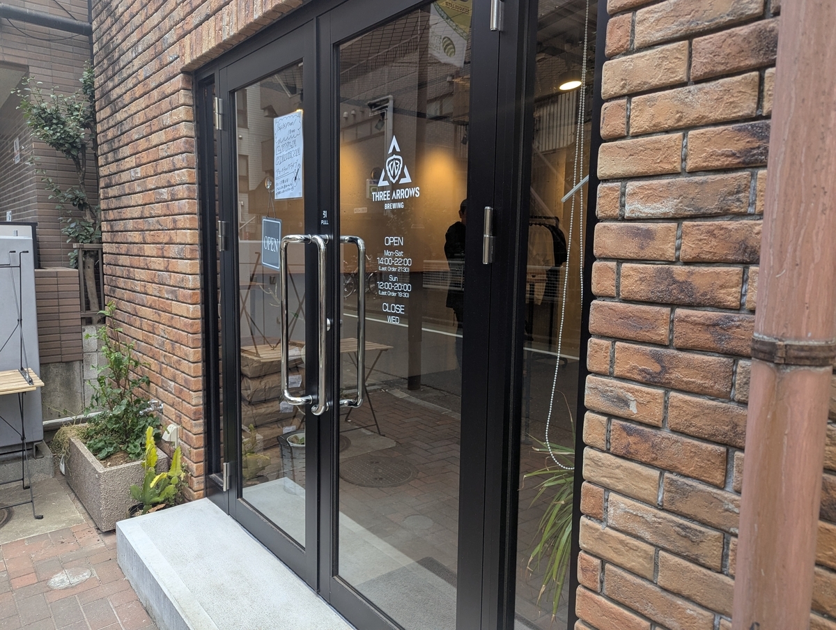 THREE ARROWS BREWINGの店舗入口。レンガ壁に囲まれたガラス扉にロゴ、営業時間（Mon-Sat 14:00-22:00、Sun 12:00-20:00、CLOSE WED）が記されている。左手にOPENの看板と植栽が見える