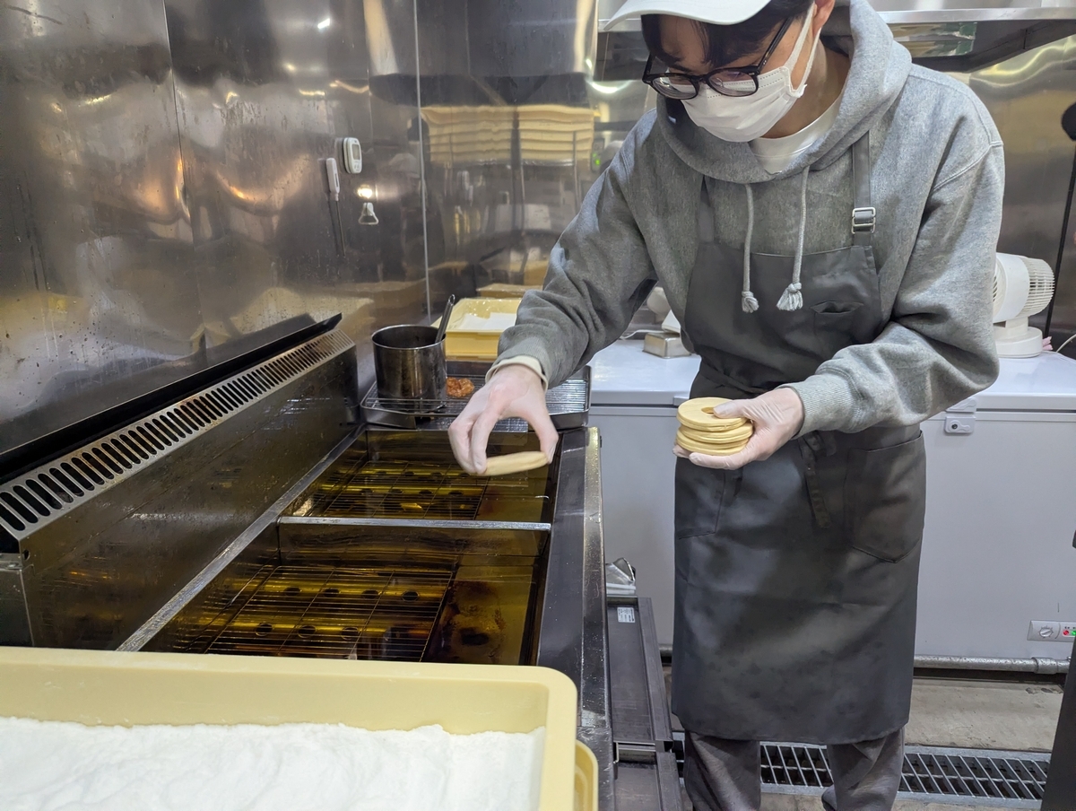 スタッフが多層に折り重なったフレーキードーナツの生地をフライヤーに投入する様子。左手に数枚重ねた薄い円形の生地を持っている