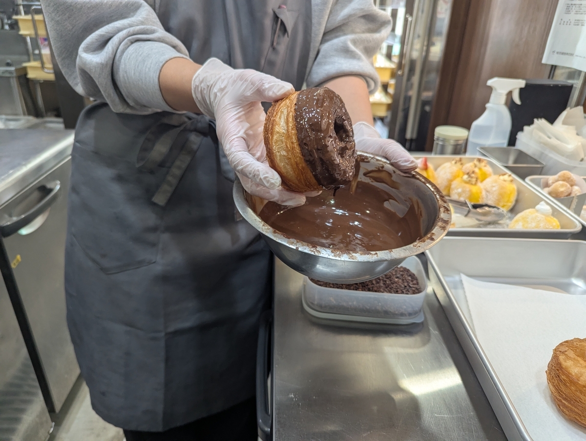 スタッフがフレーキードーナツの片面をボウルの溶かしチョコレートに浸している様子。手前にカカオニブの容器が見える