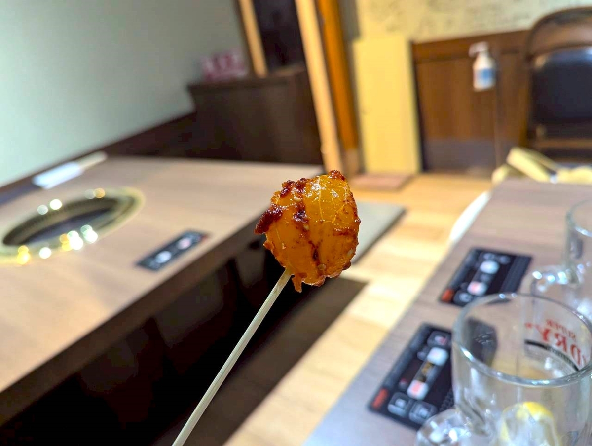 Beef Bossのみかんキムチを串で持ち上げた一枚。オレンジ色のみかんにキムチの赤い衣がまとわりつき、背景に店内のテーブル席が見える