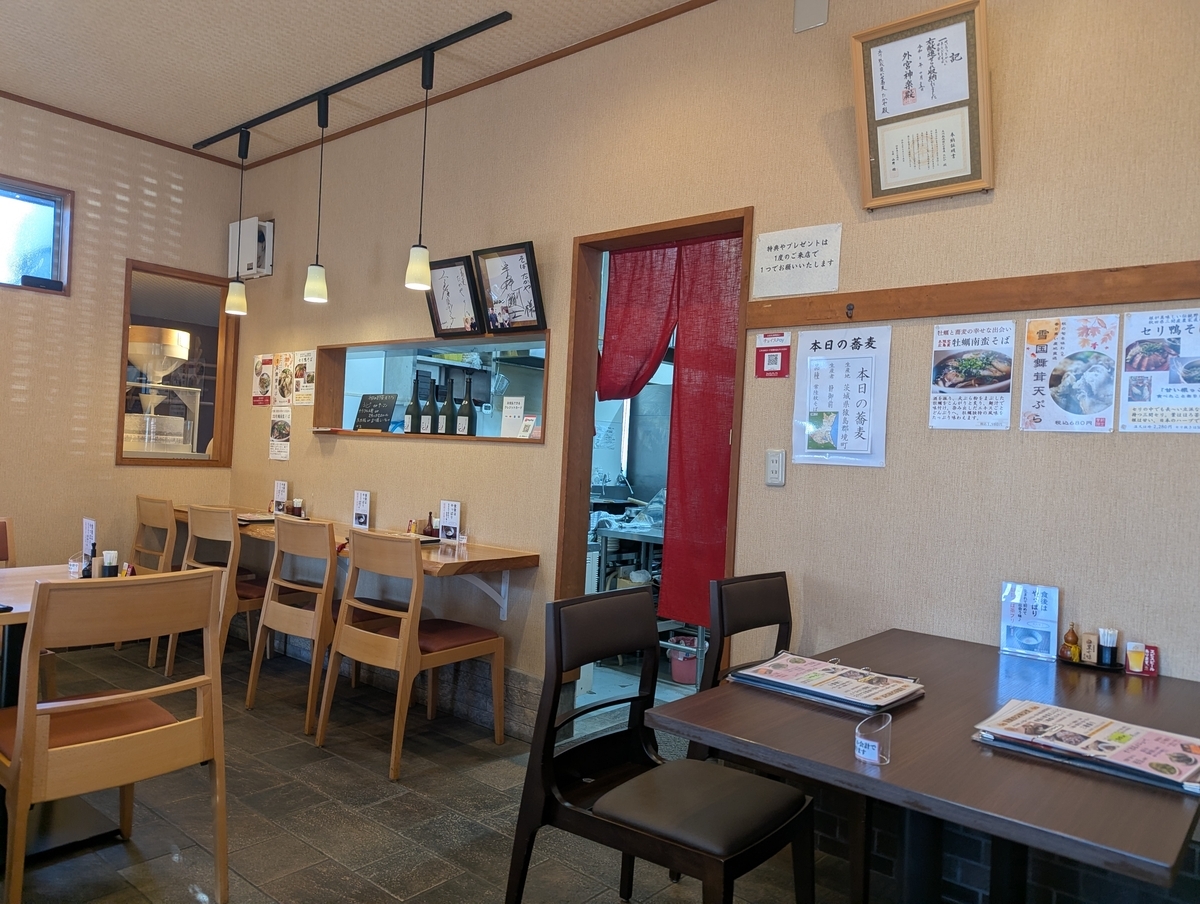 立川熟成寝かせ蕎麦たかやの店内。木のカウンター席とテーブル席が並び、奥には厨房と赤い暖簾、壁には季節メニューの貼り紙と受賞証