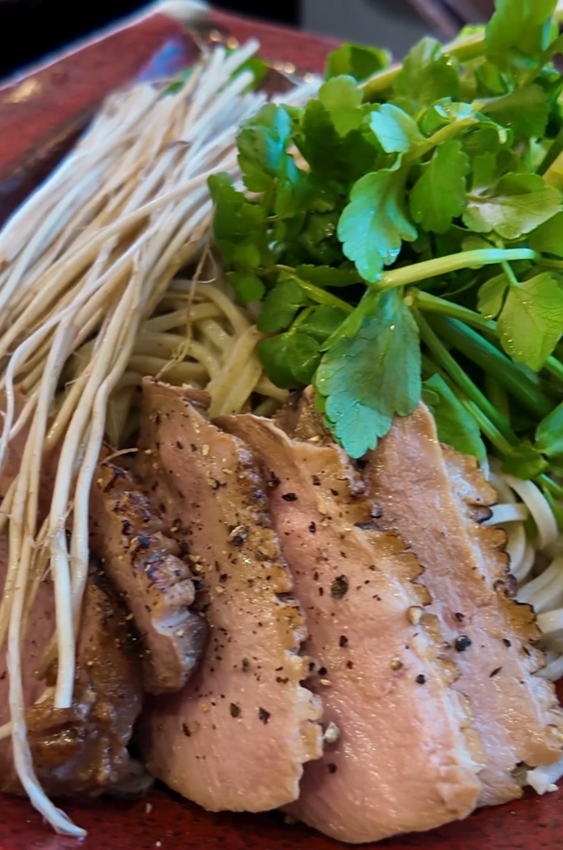 立川たかや「三関セリの鴨そば」のクローズアップ。皮目に焼き目をつけた鴨ロースのスライス、根を残した三関セリの白い長根と緑の葉、そばの麺が一枚の朱塗りの皿に盛り付けられている