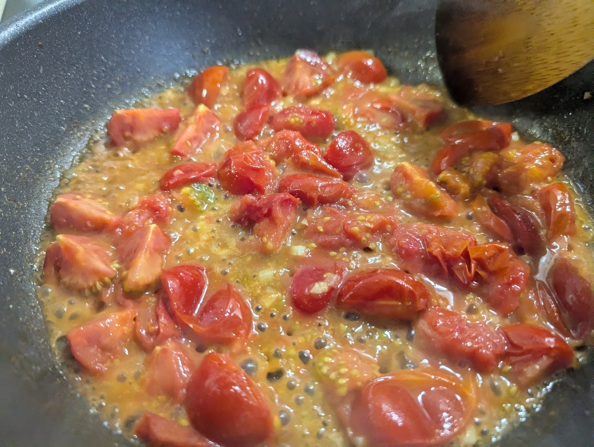 フライパンで加熱中のさわとまと。半割りにしたフルーツトマトが熱でとろけ始め、果肉から染み出した果汁とオレンジ色のソースが鍋の中で煮詰まっていく様子。木べらが添えられている