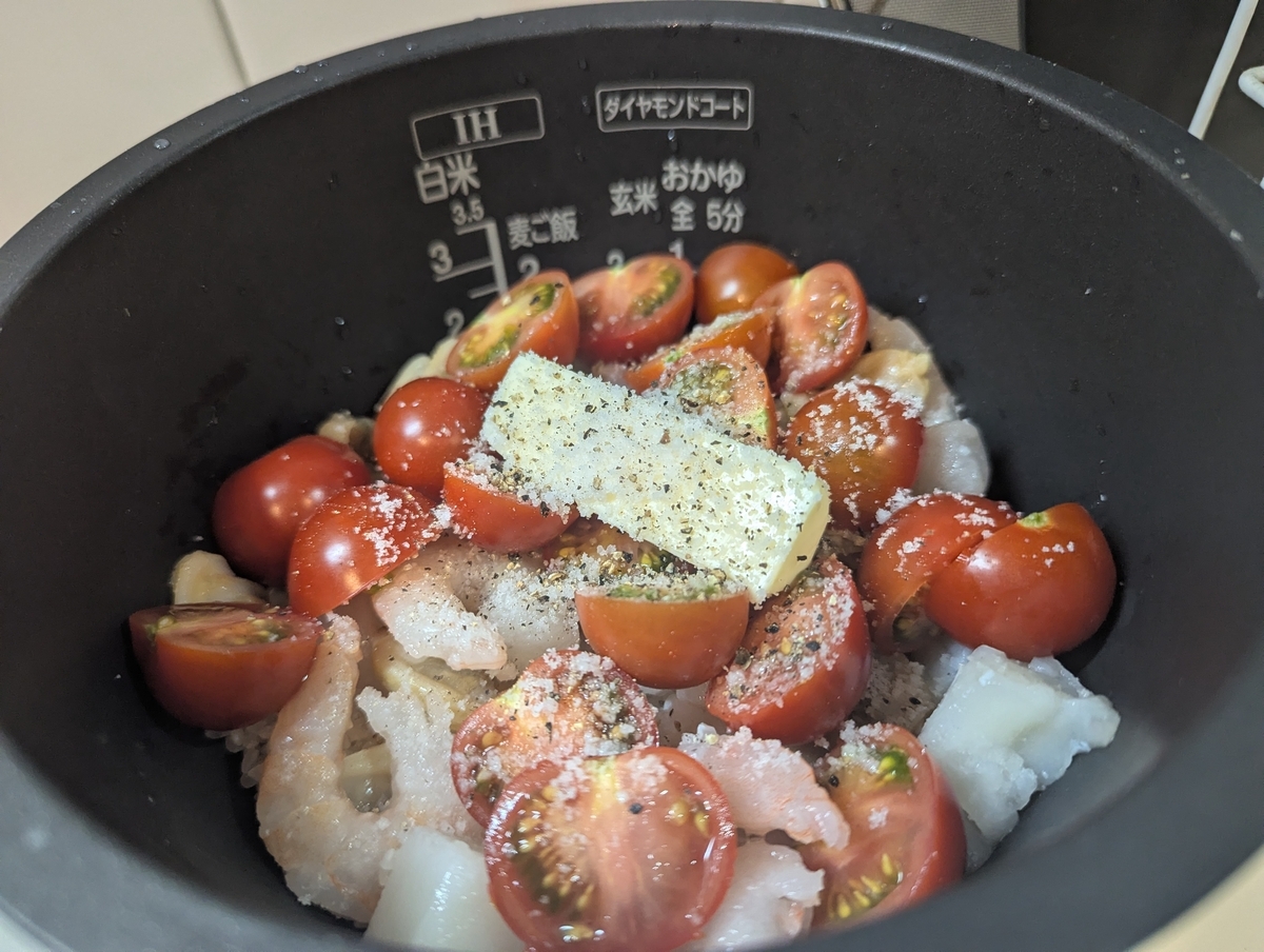 炊飯器の内釜に、半割りのさわとまと・冷凍シーフード・玉ねぎ・バター・調味料が並べられた炊飯前の仕込み状態。「IH 白米 ダイヤモンドコート」の文字も見える