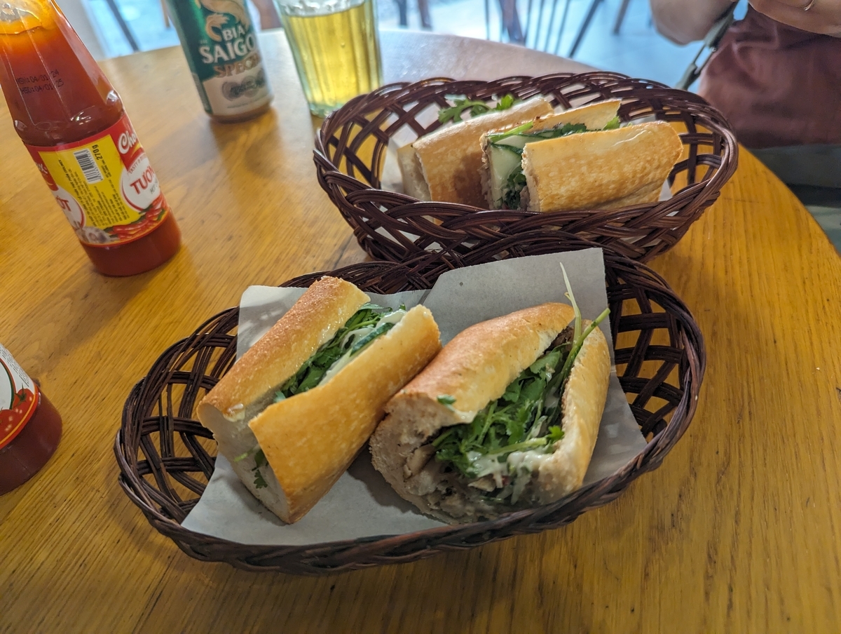 ホーチミンの現地店で食べたベトナム名物バインミー2種。バゲットに香草・肉・野菜が挟まれ、サイゴンビールとチリソースが添えられている。バナナ籠の盛り付けがベトナムらしい