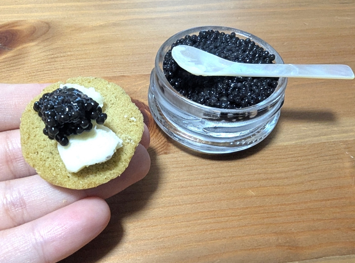 宮崎キャビア1983のキャビアと無塩バターをのせたほうじ茶ブリニを手に持つ様子。背景にシェルスプーン入りのキャビア瓶
