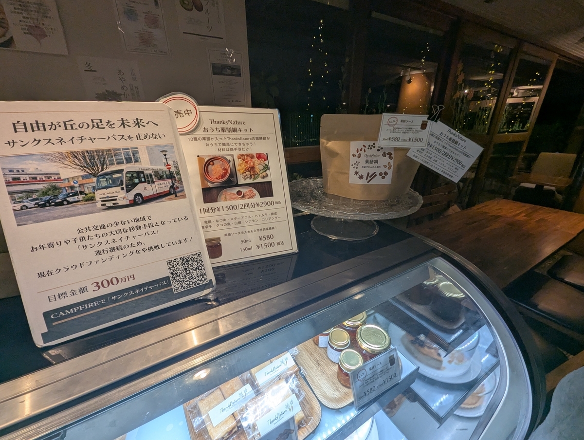 サンクスネイチャー恵比寿店内のカウンター上、自由が丘を走る送迎バス「Thanks Nature Bus」のクラウドファンディング案内POPと、おうち薬膳鍋キットの販売案内