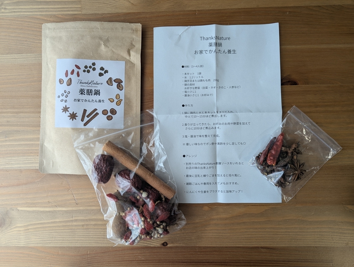 おうち薬膳鍋キットの中身一式、薬膳素材2袋、レシピ説明書、パッケージラベルが木目テーブルに並ぶ