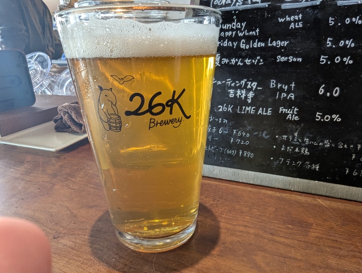 26Kブルワリーのロゴ入りグラスに注がれた黄金色のFriday Golden Lagerと、背後の黒板メニュー