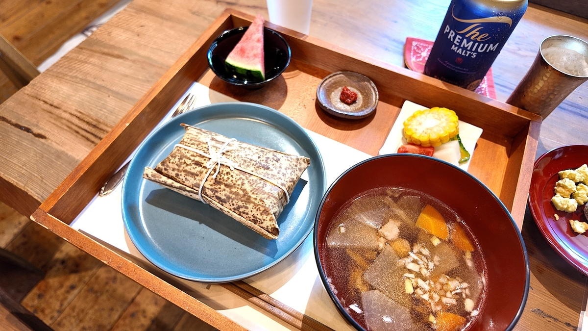 頑亭文庫のちまきセット。竹の皮に包まれたちまき、お汁、すいか、夏野菜の小皿が木のトレイに並ぶ